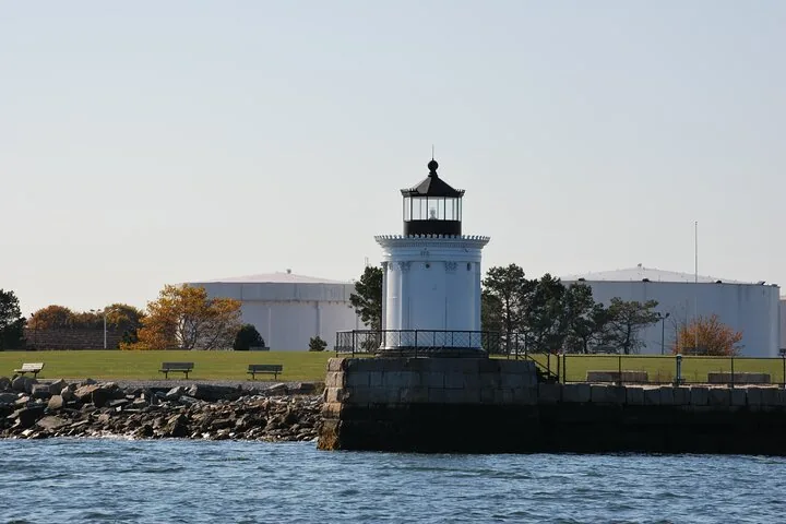 Una pequeña estructura blanca de faro se asienta sobre un muelle de piedra con grandes tanques detrás.